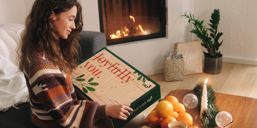 Donna apre il calendario dell'Avvento. Scritta Joyfully you. e nu3. Su un tavolo con candela, arance e decorazioni.