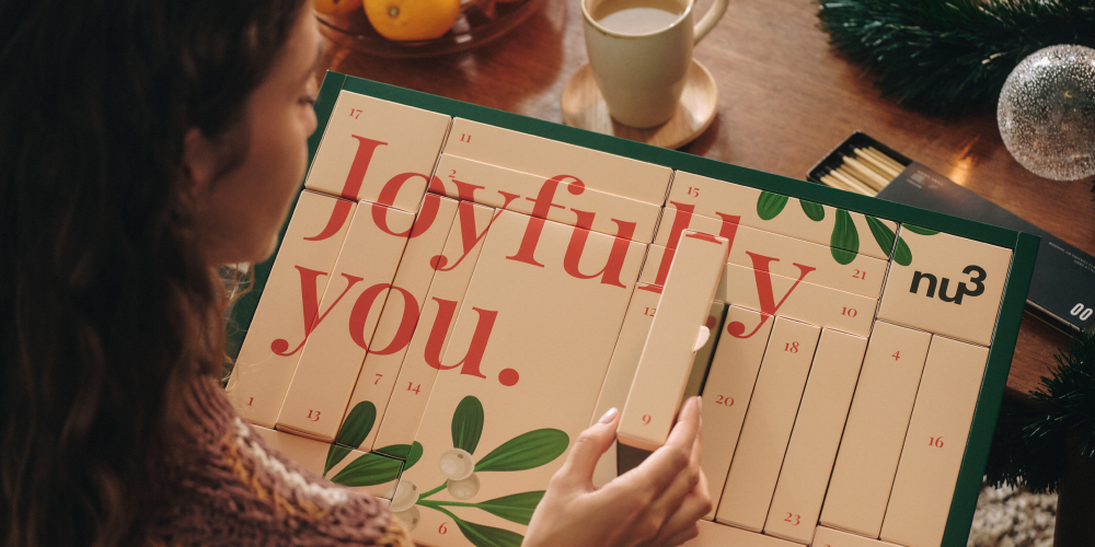 Donna apre una casella del calendario dell'Avvento. Scritta Joyfully you. e nu3. Su un tavolo con tazza e decorazioni.